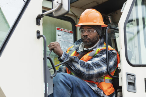 A mature African-American man working at a construction site as a crane operator, sitting in the crane cab. He is wearing protective workwear - a hardhat, safety goggles, and reflective vest.