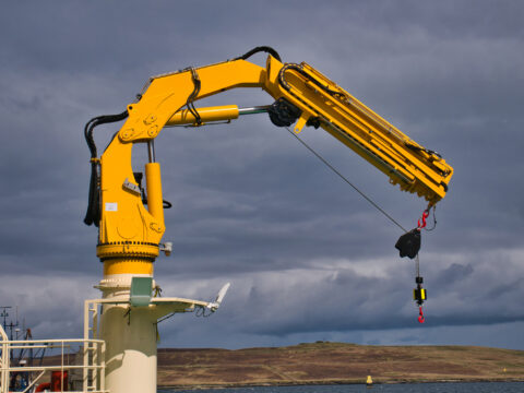 A yellow HS Marine AK 4818.5 fully foldable knuckle and telescopic boom type marine crane. Taken in sunlight with a grey sky.