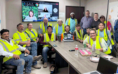 Room filled with multiple people and the majority wearing a yellow construction vest, sitting around a long conference table and in front of TV mounted on the wall